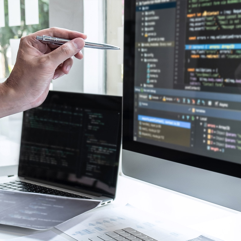 Hand eines Entwicklers zeigt mit einem Stift auf einen Monitor mit Code, während weitere Bildschirme mit Programmieroberflächen sichtbar sind.