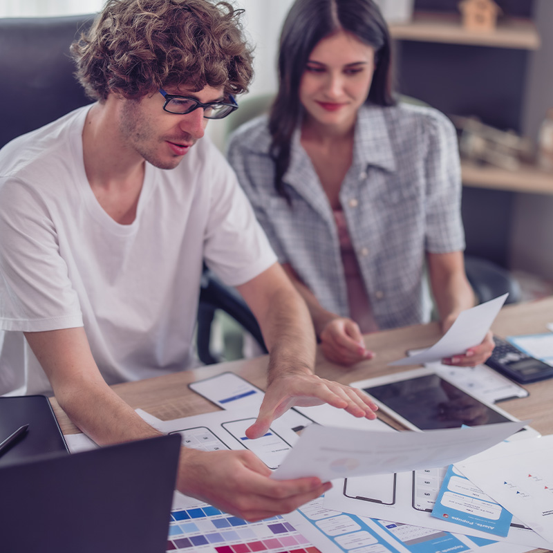 Zwei IT-Freelancer arbeiten an einem Designprojekt mit Skizzen und Farbschemata auf einem Schreibtisch.