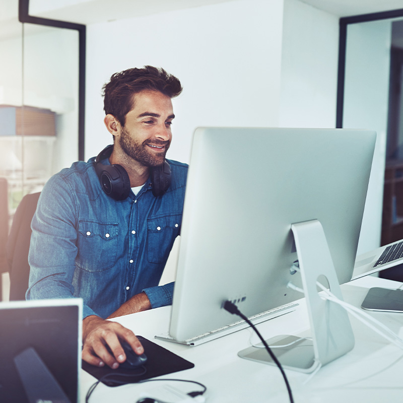 Lächelnder IT-Freelancer mit Kopfhörern um den Hals arbeitet an einem Computer in einem modernen Büro.
